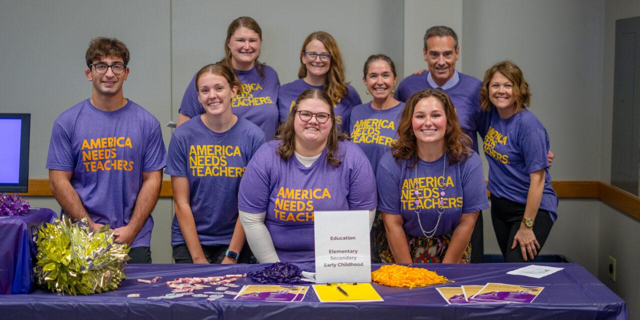 Group of people wearing purple shirts with yellow writing "America Needs Teachers"
