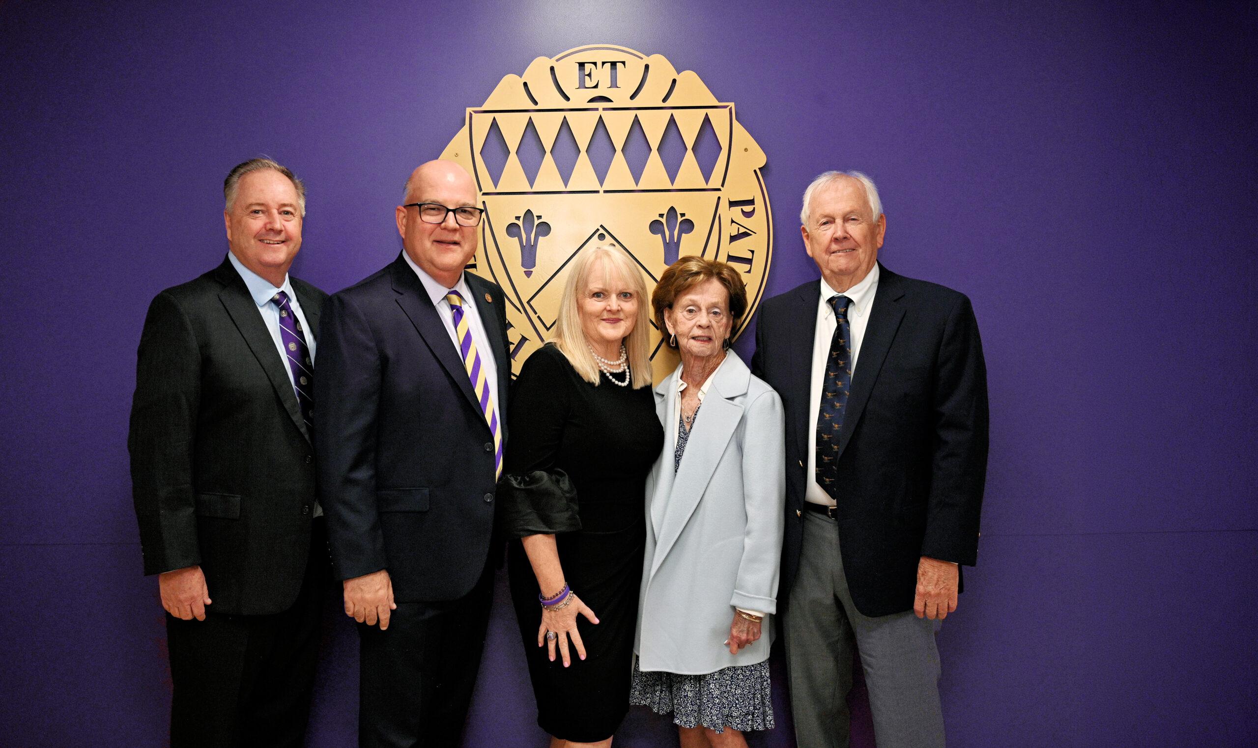 Regent Mike Rice II, Regent Emeritus Mike Rice ('64) and Geri Rice, Loras College President Mike Doyle, and Martha Doyle pose for a photo in front of a purple wall featuring the Loras College crest. The group is dressed in formal attire and stands together during a Loras College event.