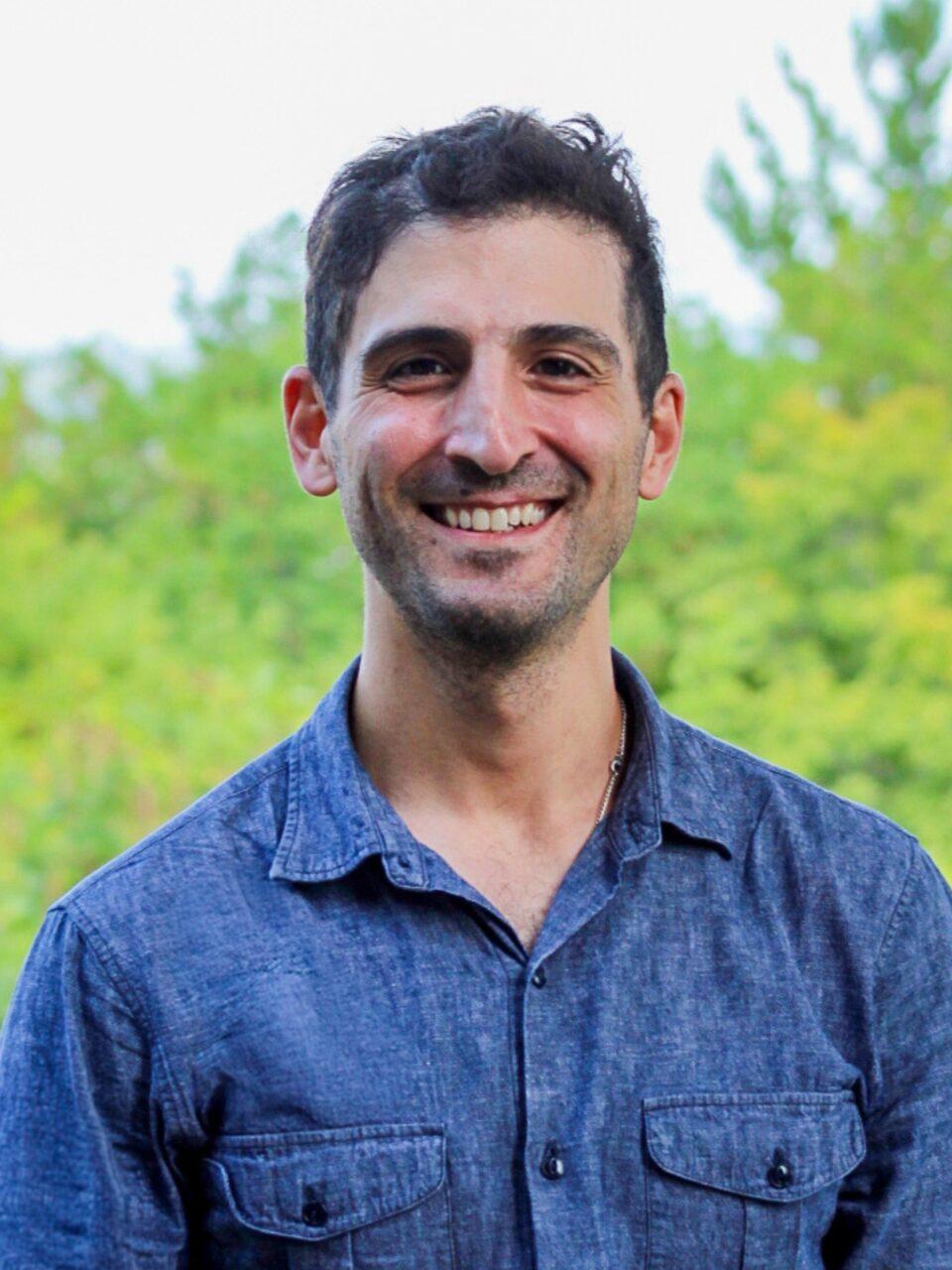 Employee wearing a blue shirt and standing in front of green trees