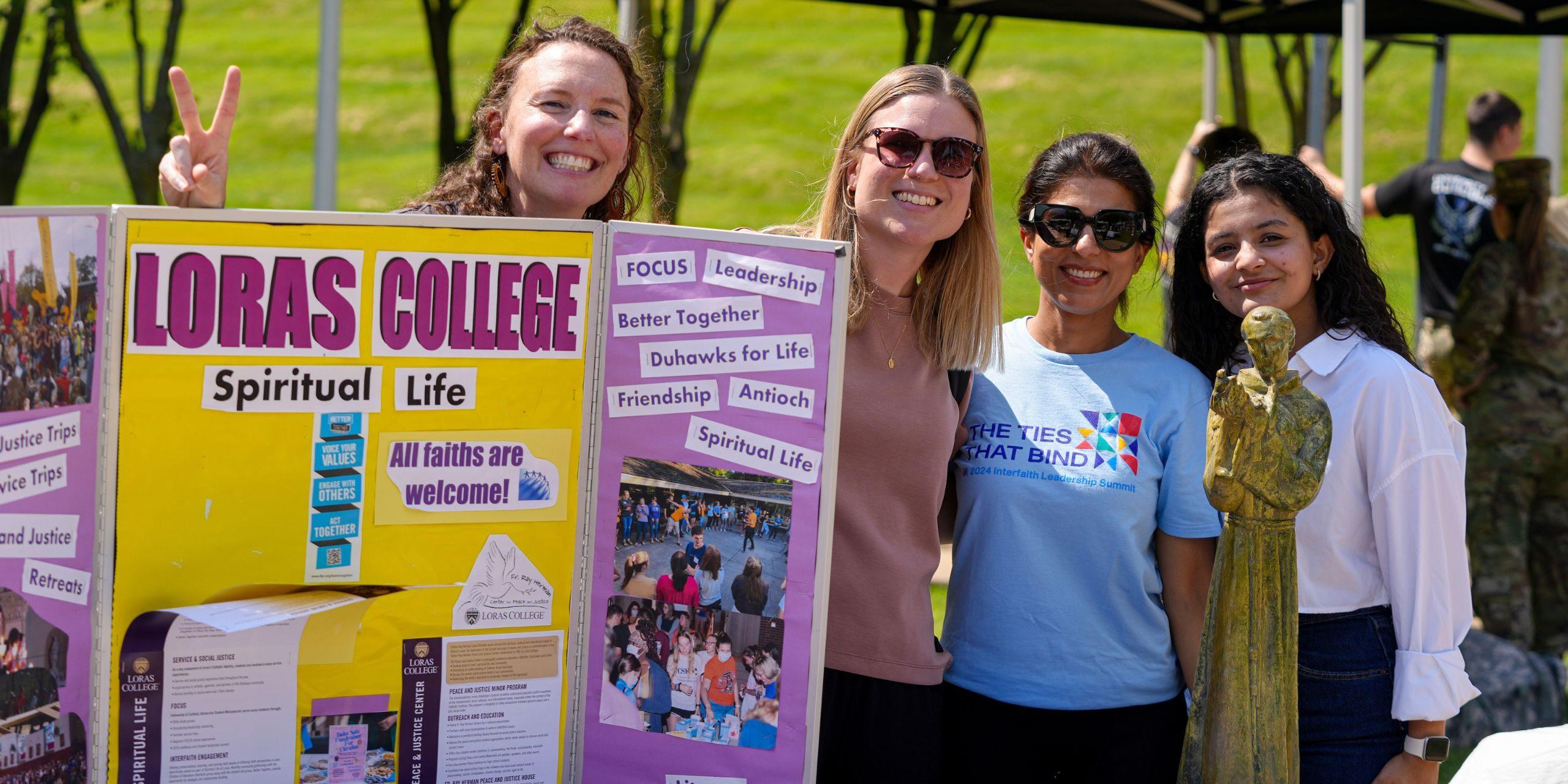 four people standing by purple and yellow board with "Loras College Spiritual Life" in big Purple letters