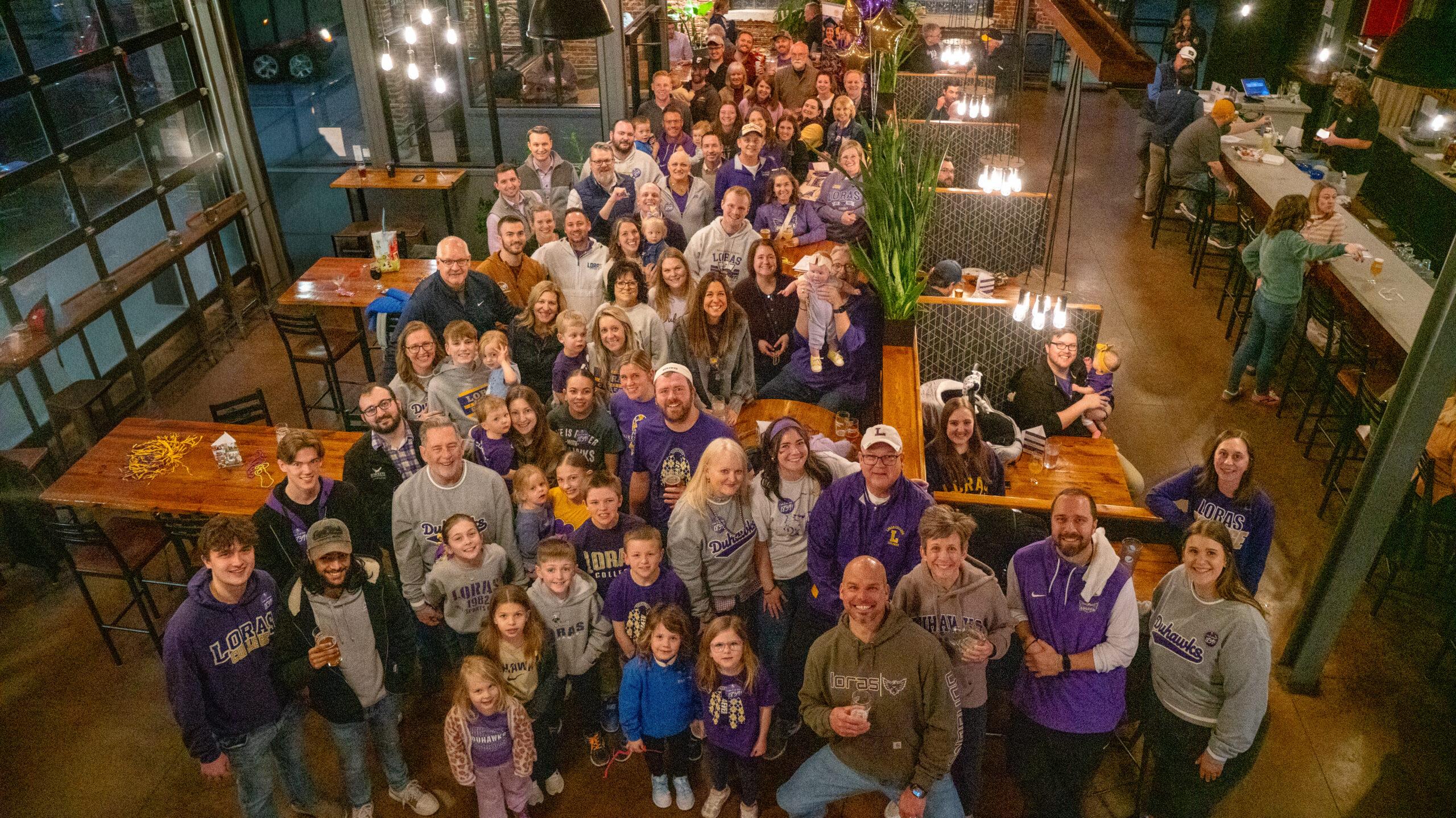 A large group of Loras College alumni, families, and supporters gathered inside a modern brewery for a Duhawk Day celebration, many wearing purple and gold Loras apparel. Adults and children stand closely together smiling toward the camera, surrounded by wooden tables, hanging lights, and a lively indoor setting.