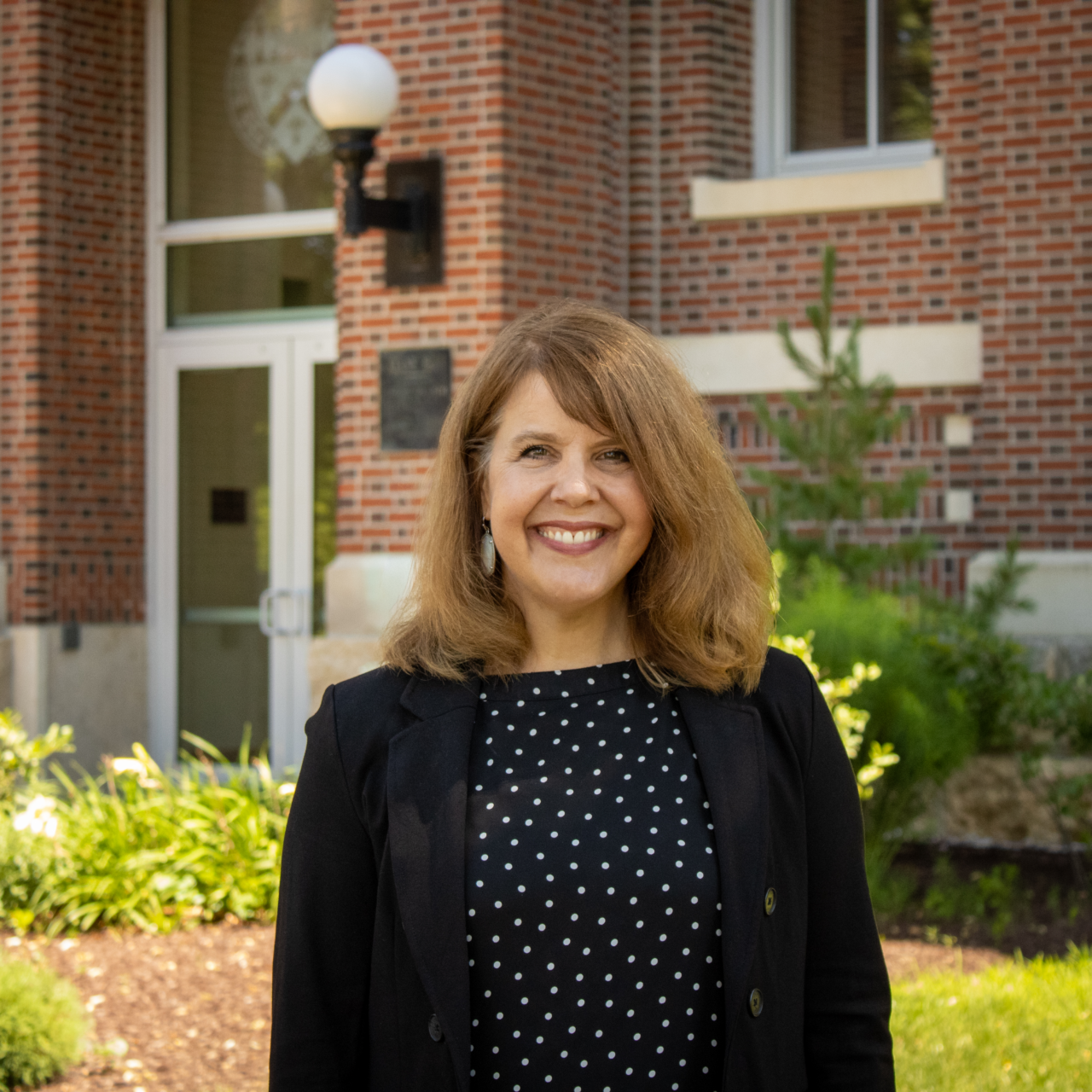 Woman smiling in front of a brick building wearing a black and white polka dot blouse and a black blazer