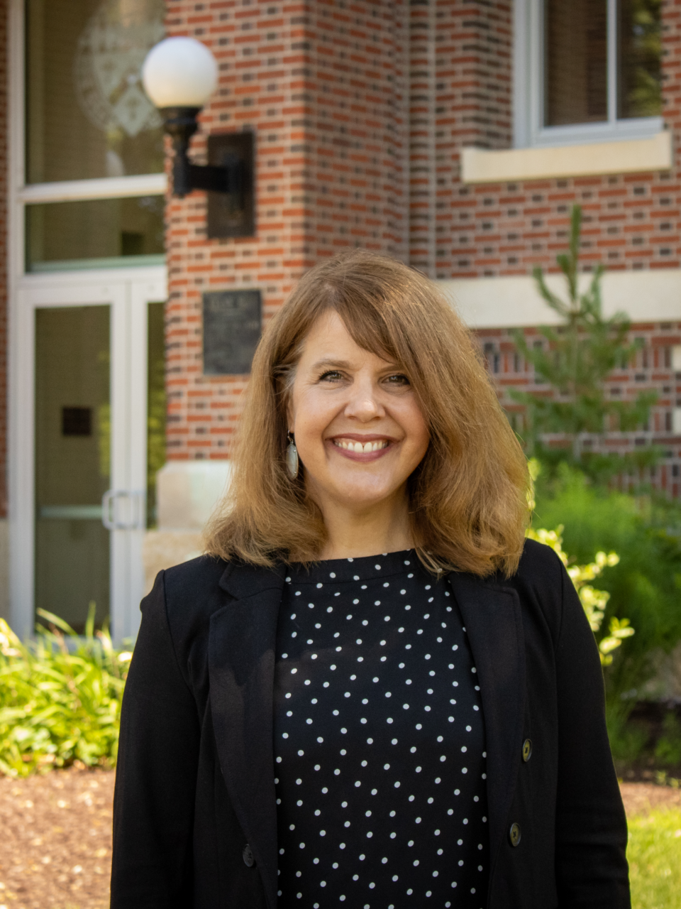 Woman smiling in front of a brick building wearing a black and white polka dot blouse and a black blazer