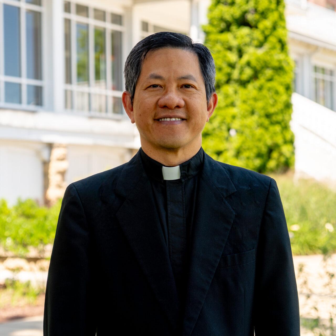 Catholic priest in a black suit with hot collar standing in front of a white porch and green trees