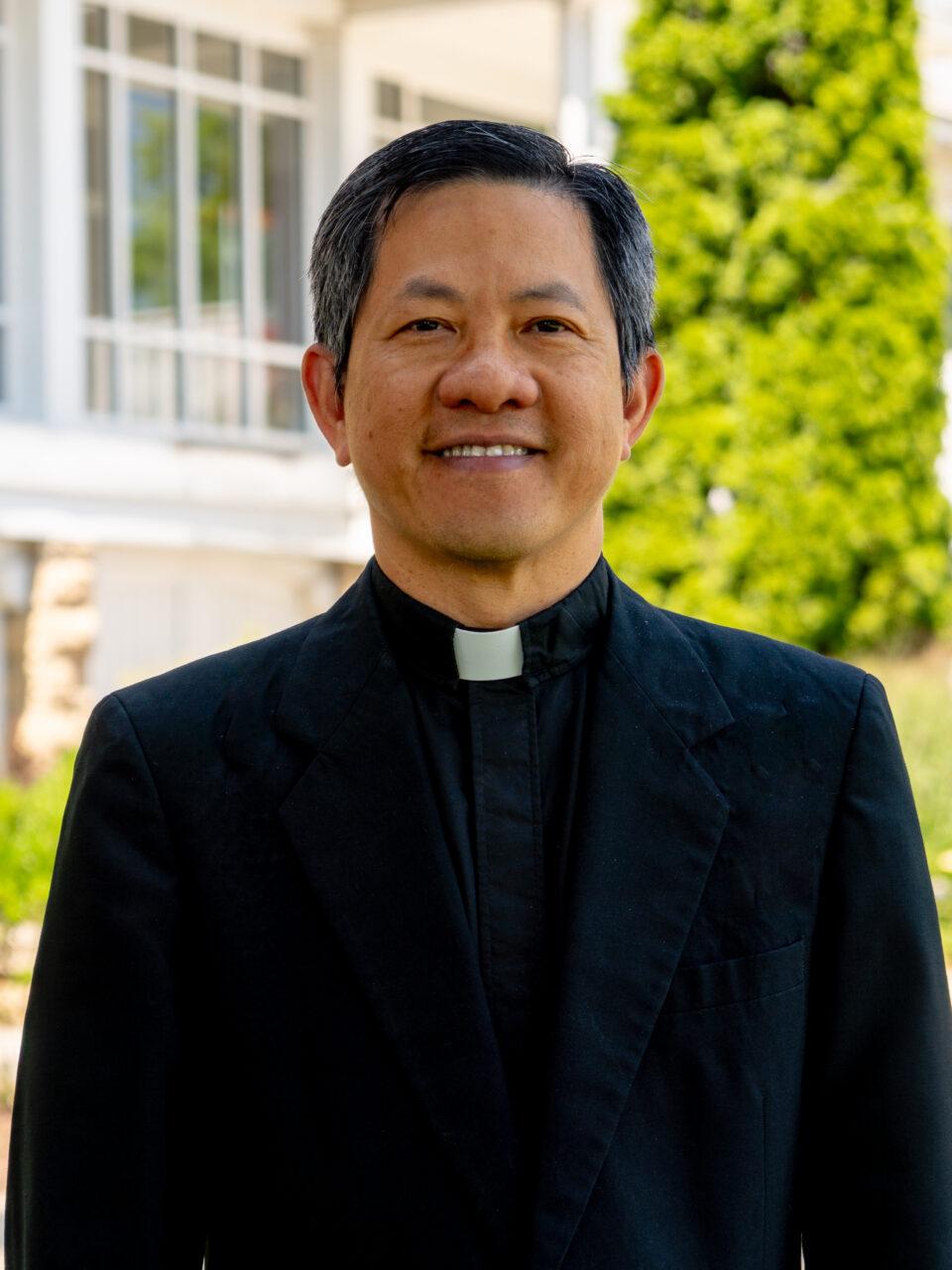 Catholic priest in a black suit with hot collar standing in front of a white porch and green trees