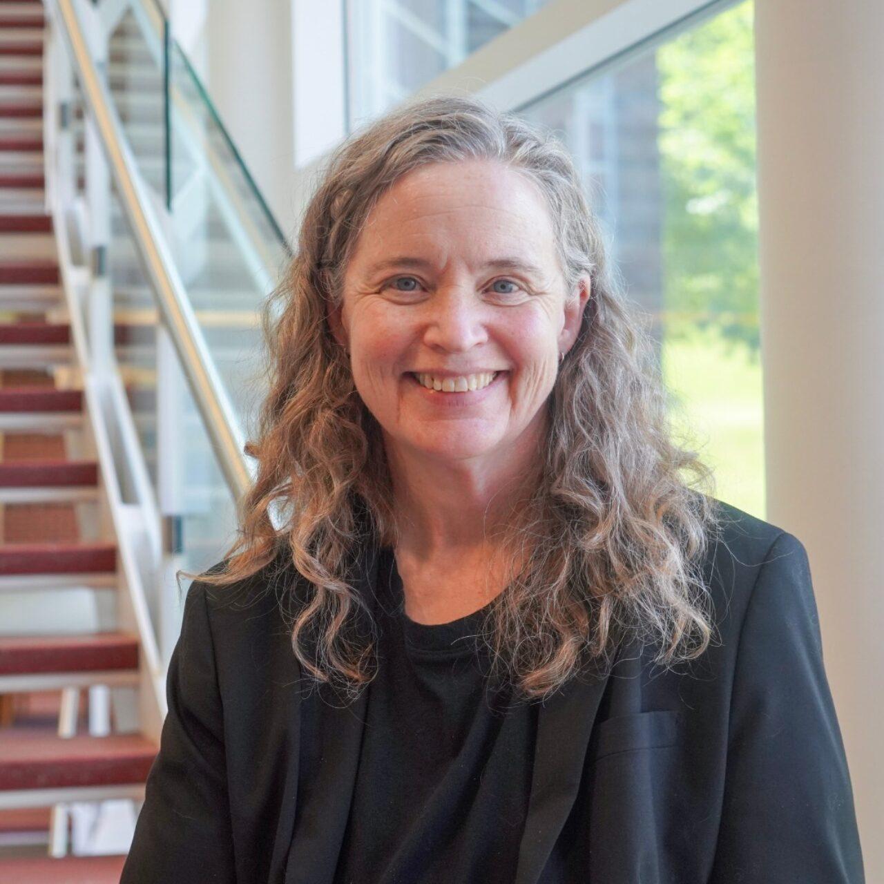 smiling employee with long hair, black shirt, and black blazer standing in front of a set of stairs