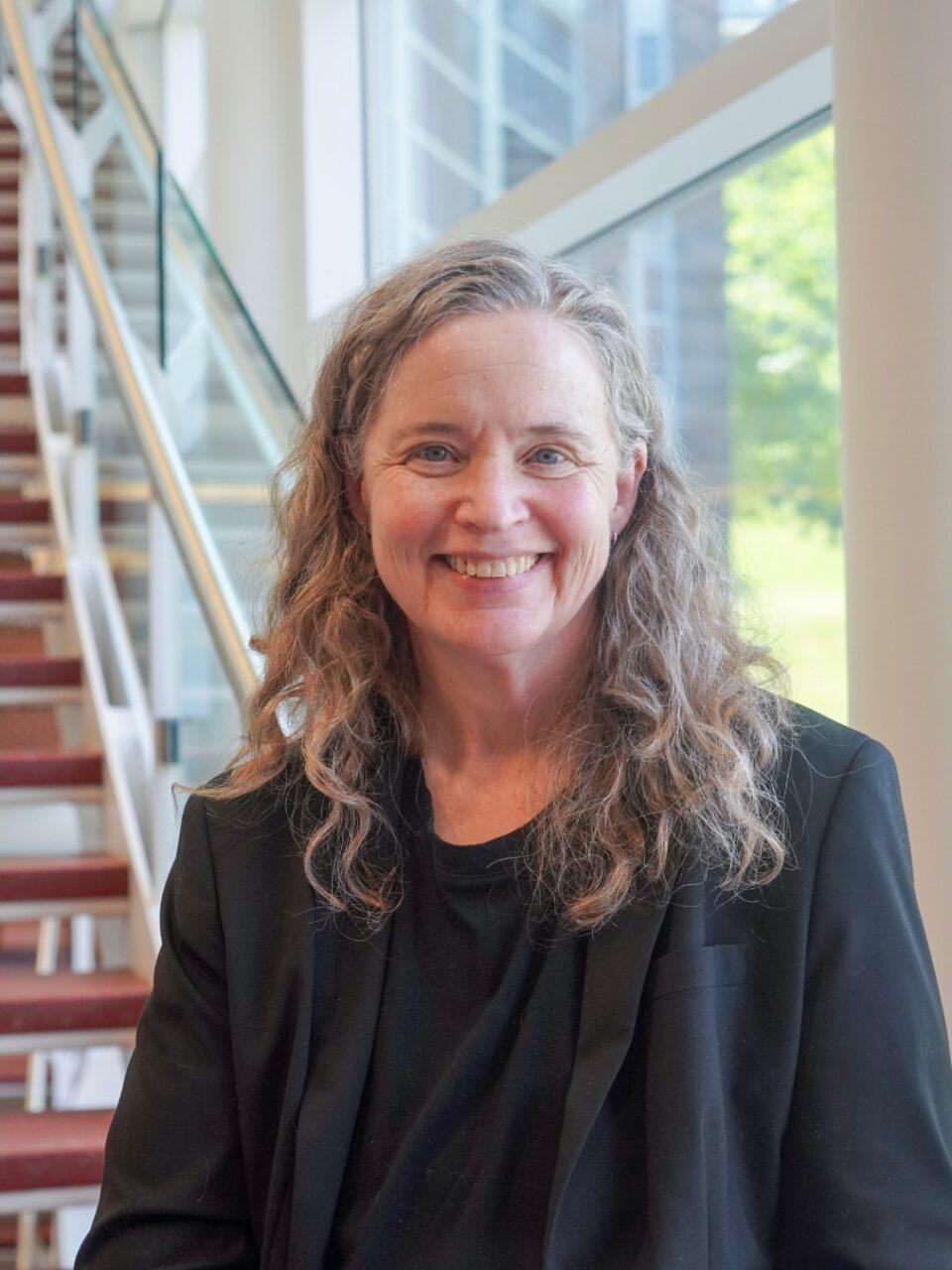 smiling employee with long hair, black shirt, and black blazer standing in front of a set of stairs
