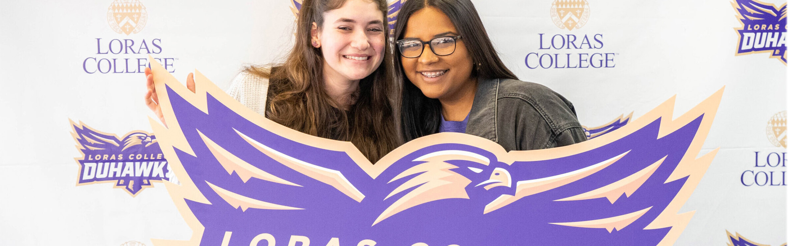 two students smiling with duhawk pride while holding up banner with the Duhawk Athletics Logo