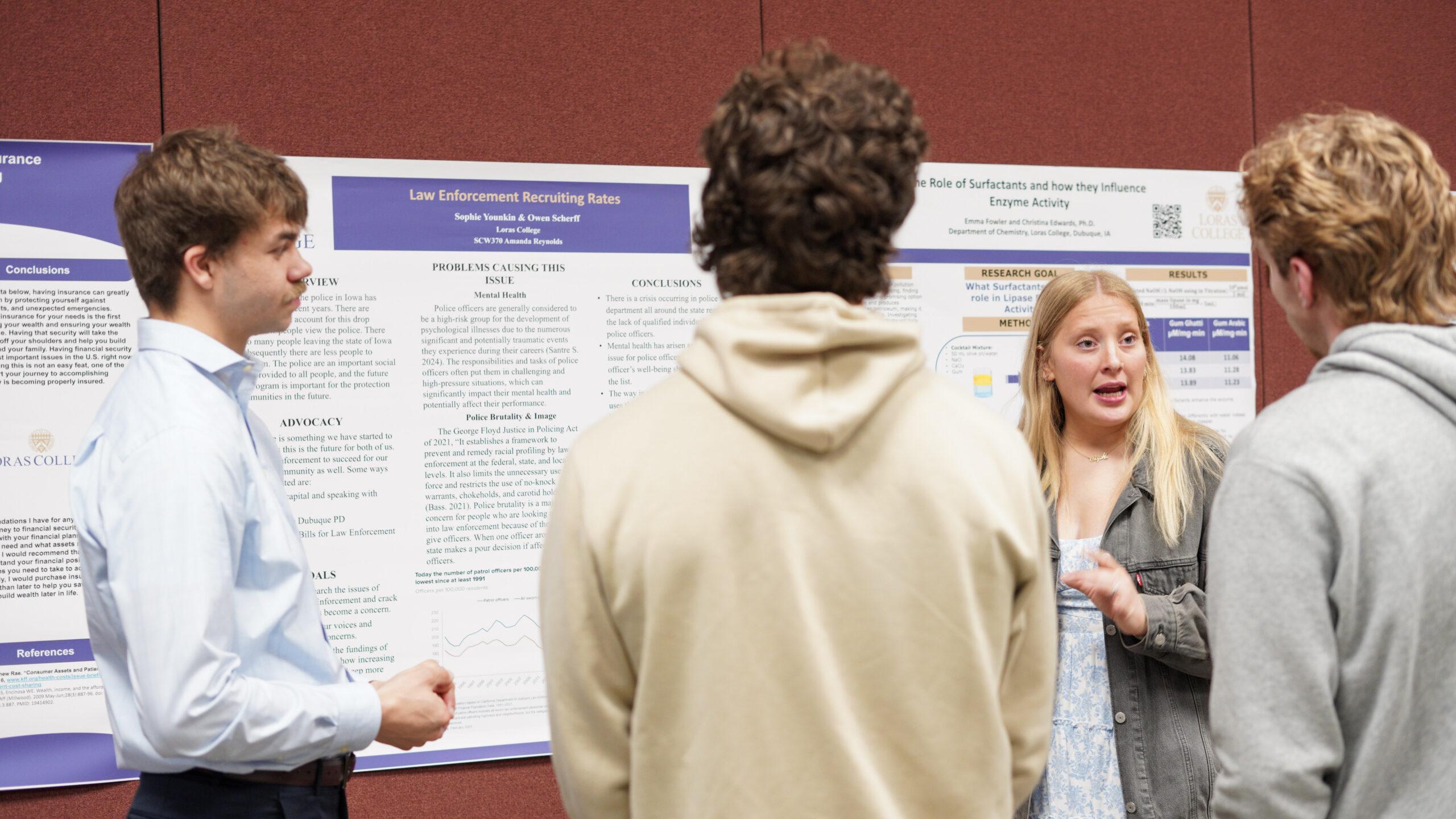 Four Loras College students stand in front of academic research posters during the Legacy Symposium, as one student presents and gestures while explaining her project to three peers in a campus event space.