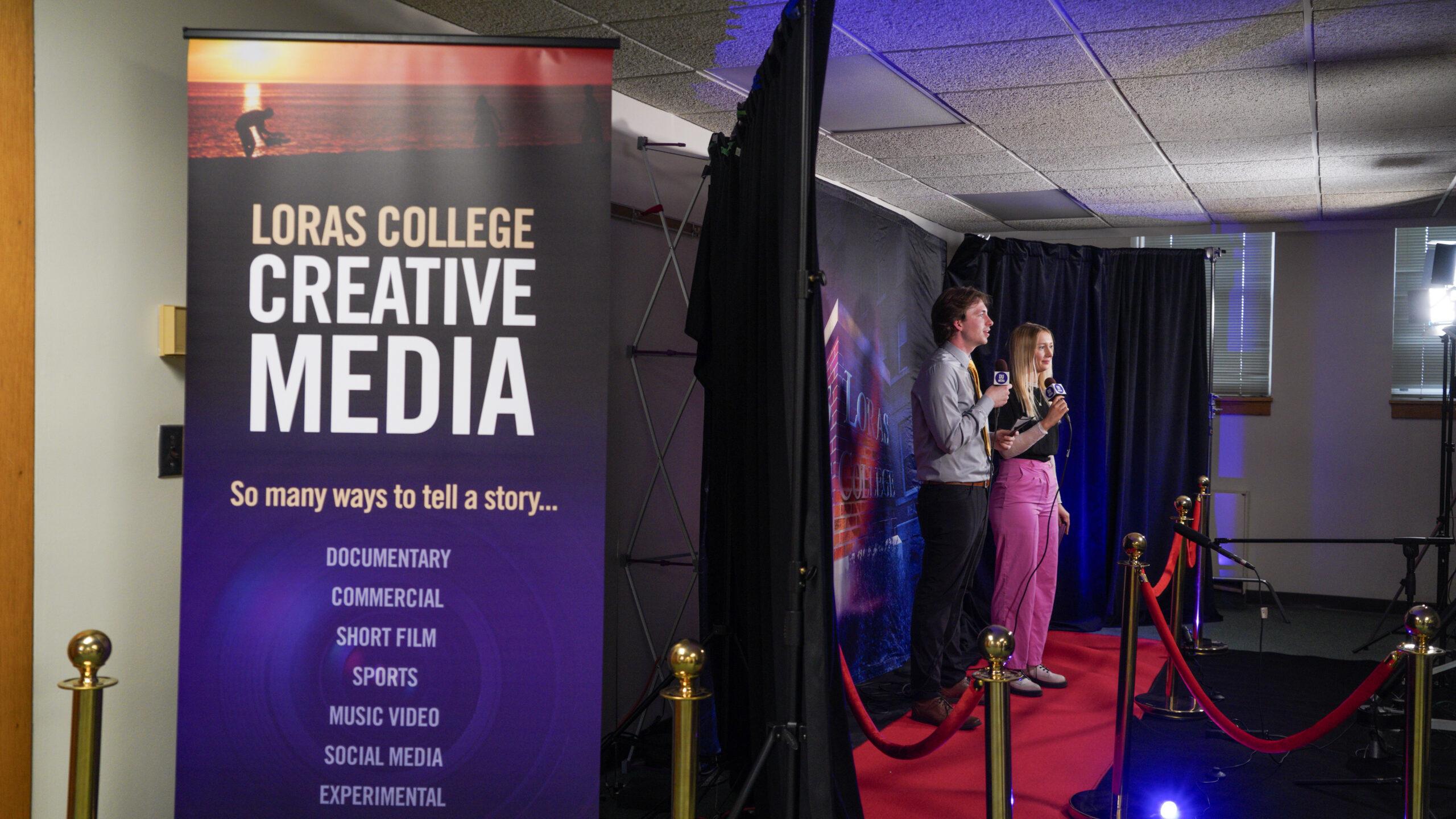 Two students stand on a small red carpet set, holding microphones and speaking on camera in a studio setup. A large Loras College Creative Media banner is displayed beside them, highlighting storytelling areas like documentary, commercial, sports and social media, with lighting equipment and backdrop visible behind the scenes.