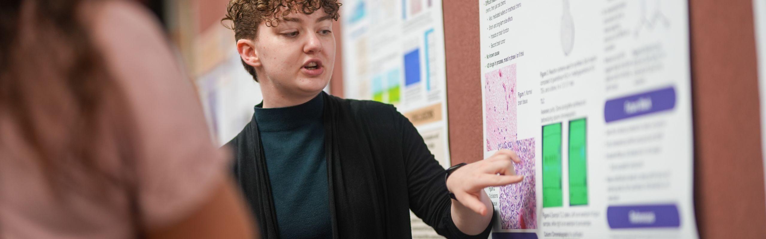 Student wearing black and green sweater explaining contents of poster.