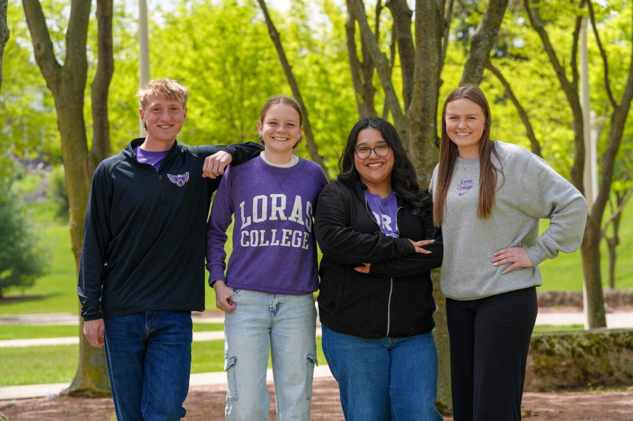 four students smiling in front of green trees in Loras College clothing