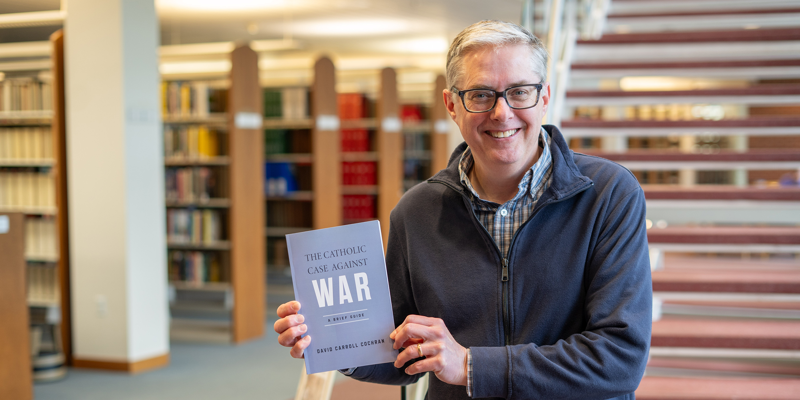 Professor David Cochran stands in the Loras College library, holding and displaying his new book, "The Catholic Case Against War: A Brief Guide."