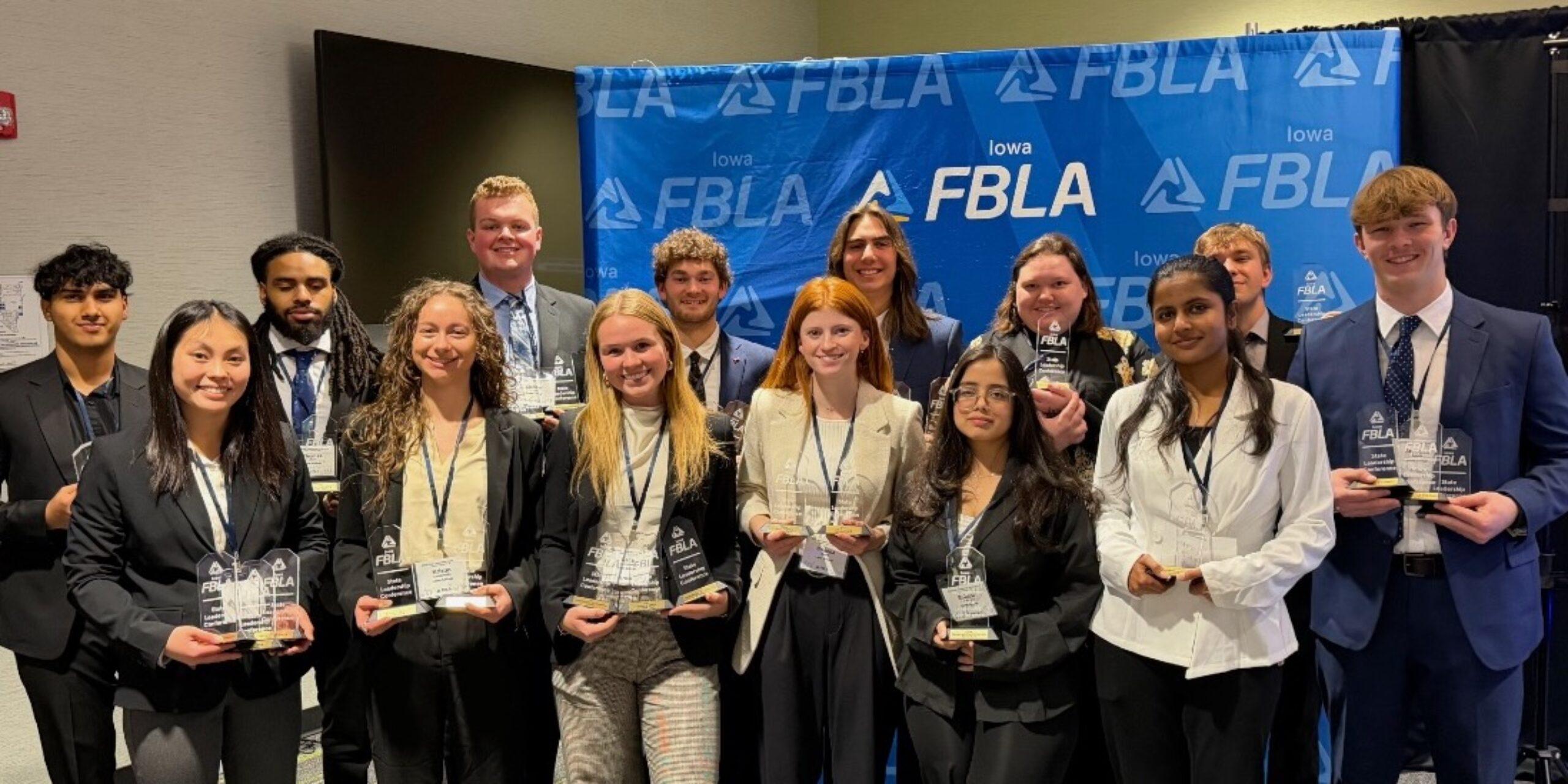 A group of Loras College students dressed in business attire pose together in front of a blue Iowa FBLA backdrop, holding multiple award plaques and smiling after a successful competition.
