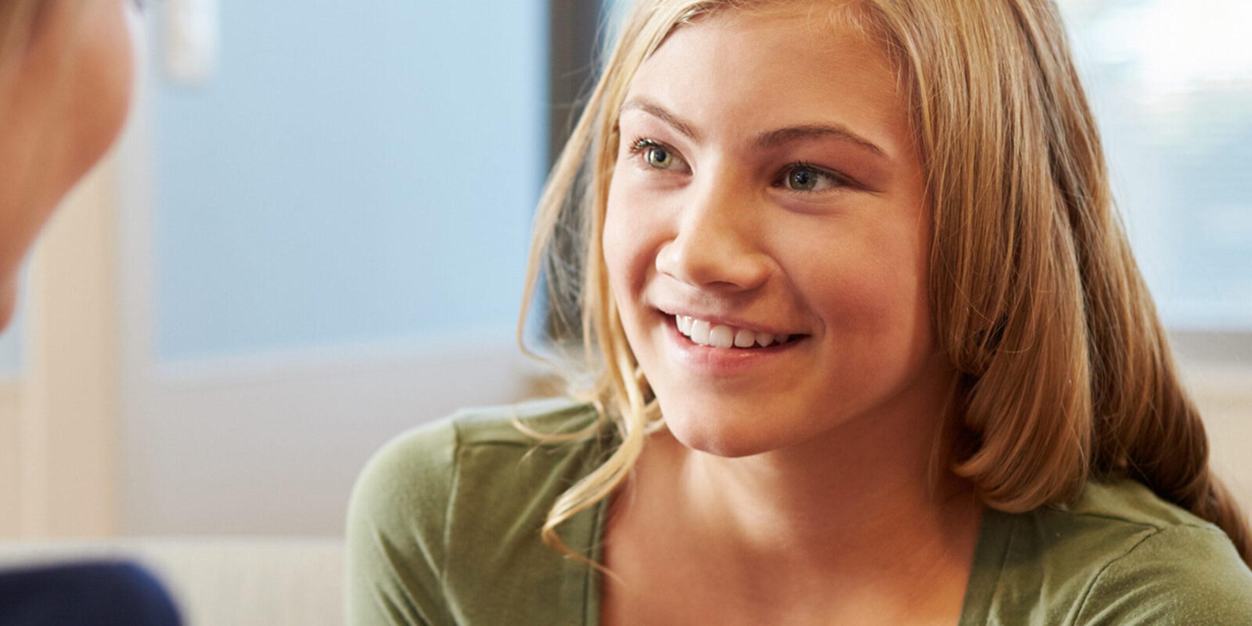 A smiling young woman with long blonde hair and a green shirt sits facing another person, engaged in a friendly conversation in a bright indoor setting.